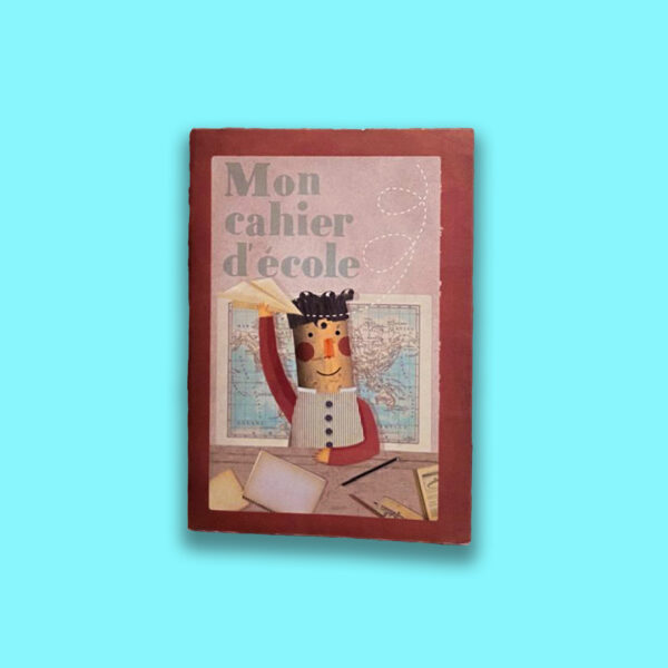 Taccuino illustrato con copertina rosa antico e scritta “Mon cahier d’école”, raffigurante un bambino che lancia un aeroplanino di carta davanti a una mappa geografica. Prodotto a Barcellona.